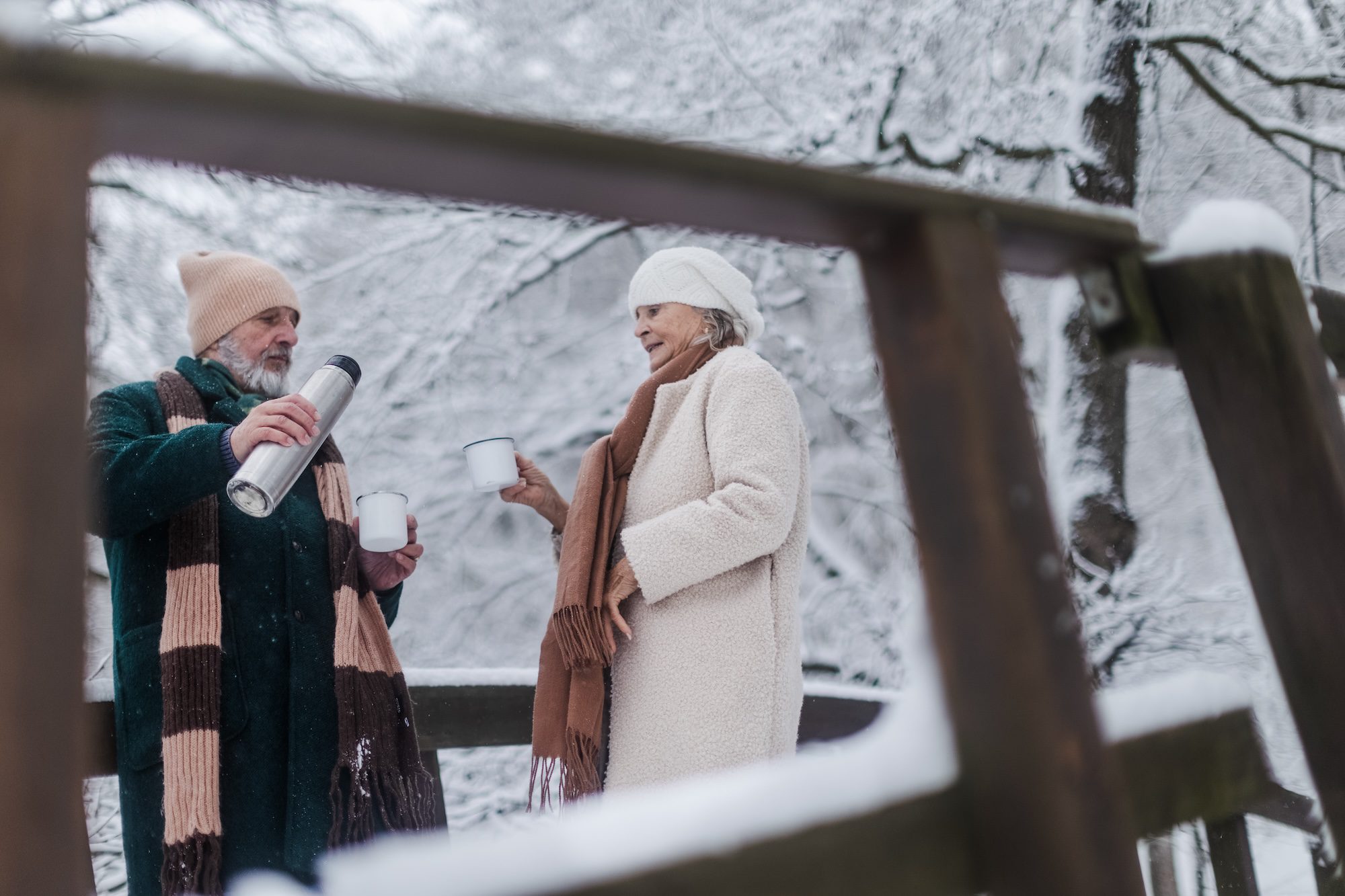 Elegant senior couple having hot tea outdoor, during cold winter snowy day. Winter vacation in the mountains. Wintry landscape.