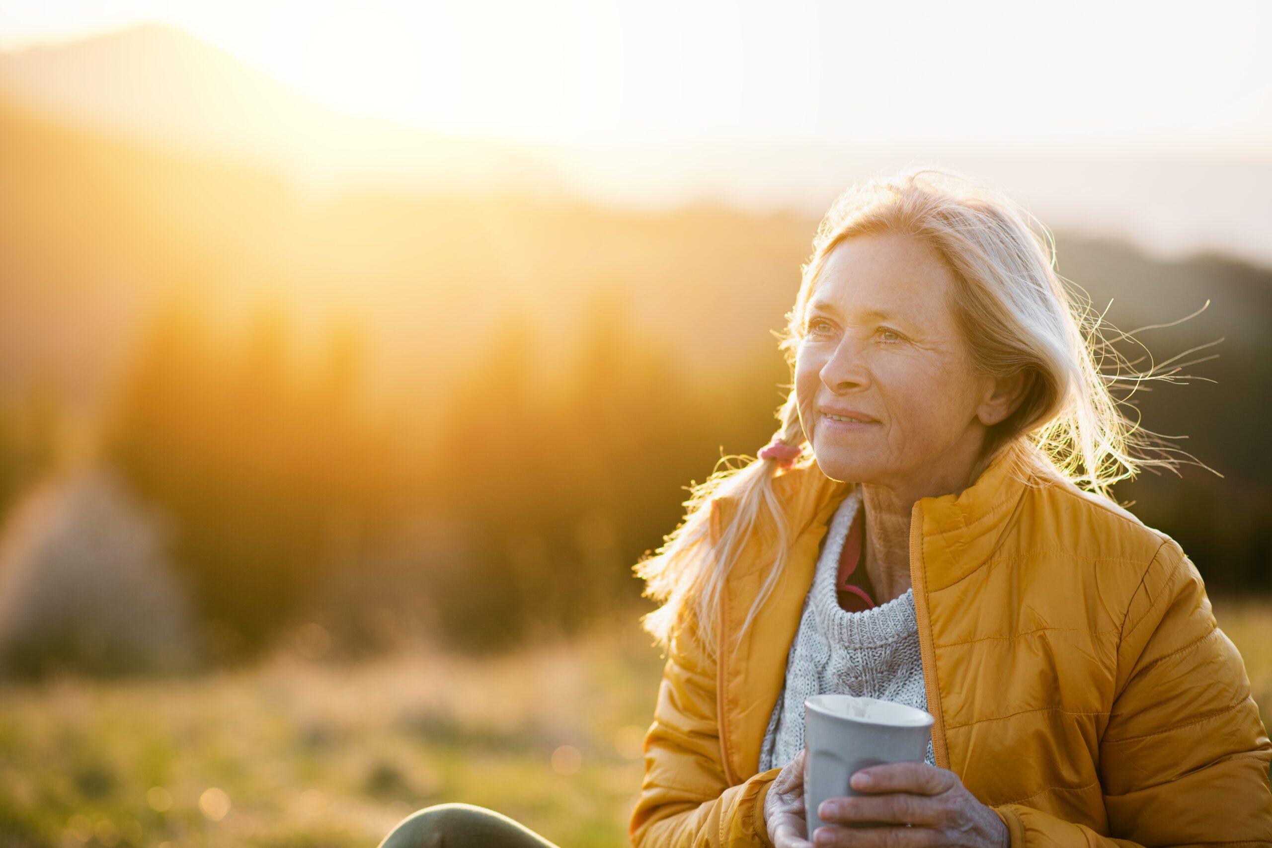 Attractive senior woman sitting outdoors in nature at sunset, relaxing with coffee.